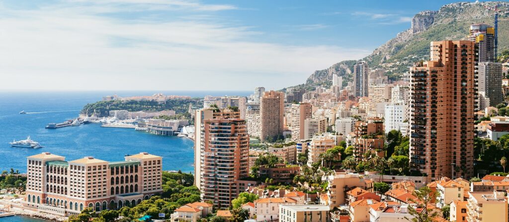 A panoramic view of Monaco featuring tall modern buildings, luxury hotels, the Mediterranean Sea with yachts, and mountains in the background beneath a blue sky with light clouds.