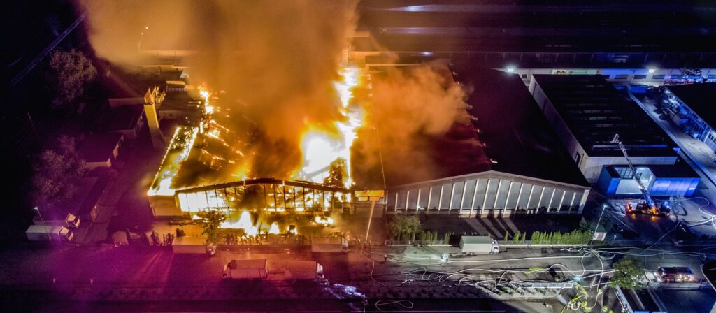 Aerial view of a large industrial building engulfed in flames at night, with thick smoke rising and emergency vehicles with hoses responding to the fire surrounding the scene.