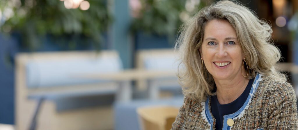 A woman with wavy blonde hair, wearing a tweed jacket and blue blouse, smiles while sitting indoors. The background is softly blurred with seating and green plants visible.
