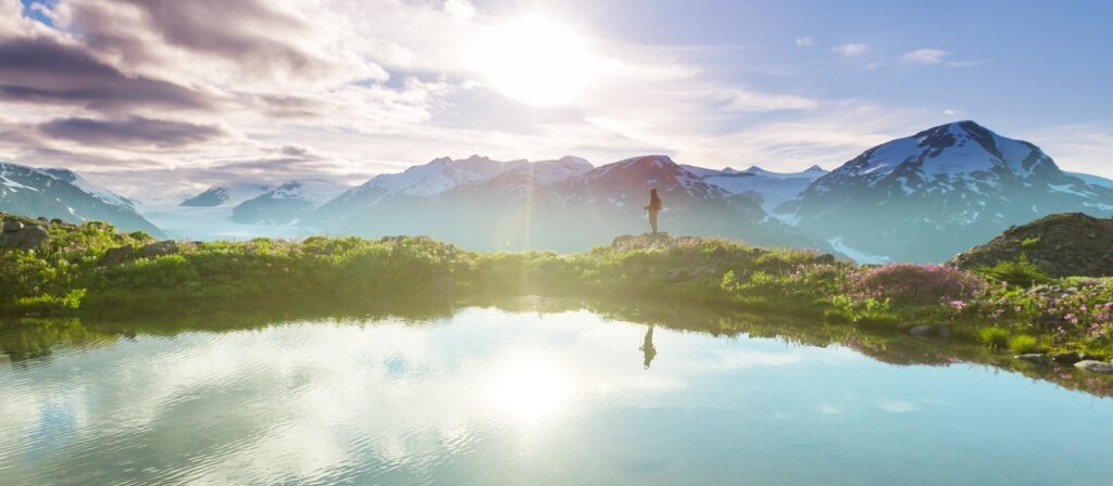 A person stands on a grassy hill near a calm lake, with snow-capped mountains and a bright sun in the background. The scene is reflected in the water, creating a peaceful and scenic landscape.