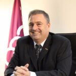 A man in a dark suit and tie sits at a desk, smiling at the camera. There is a maroon and white flag in the background and he appears to be in an office setting.