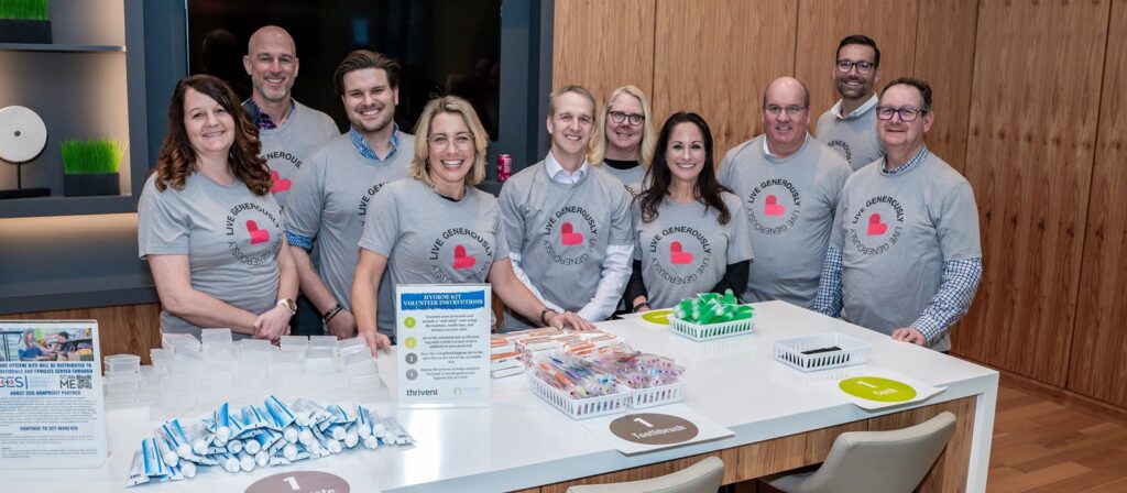 A group of ten smiling people wearing matching gray shirts with a red heart logo stand behind a table with hygiene kits, toiletries, and supplies in a modern room, appearing to volunteer for a community service event.