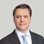 A man with short brown hair wearing a dark suit, white shirt, and light blue tie, posing against a plain light gray background.