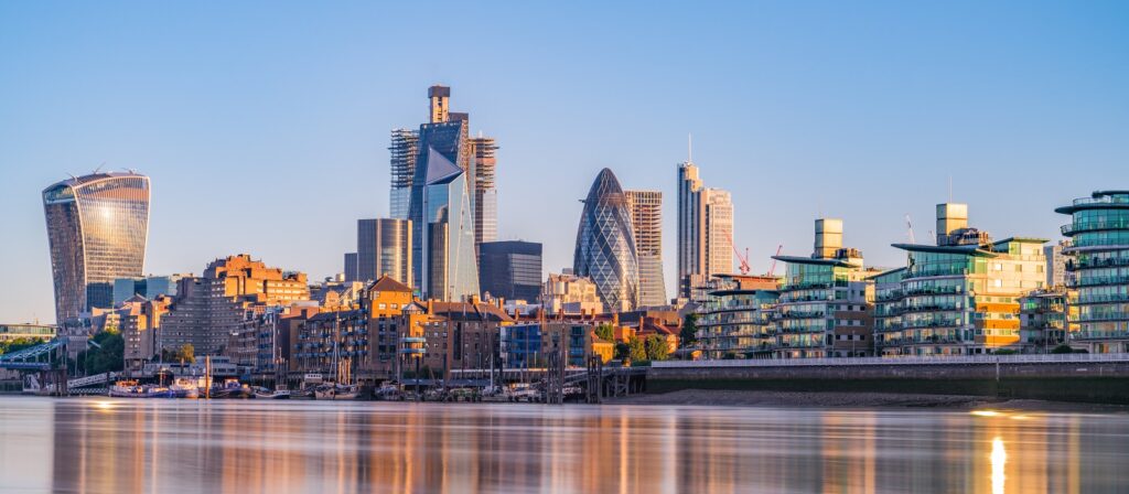 London skyline at sunrise with modern skyscrapers and historic buildings along the River Thames, including the Walkie Talkie building, the Gherkin, and glass-fronted apartments reflecting in the water.