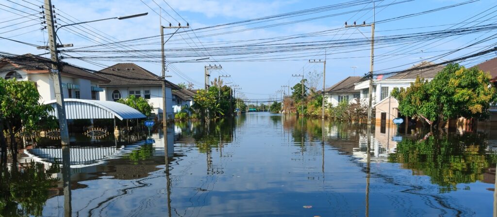 A residential street is heavily flooded, with water reaching halfway up houses. Power lines and trees line the road, and the sky is clear and blue above the flooded area.