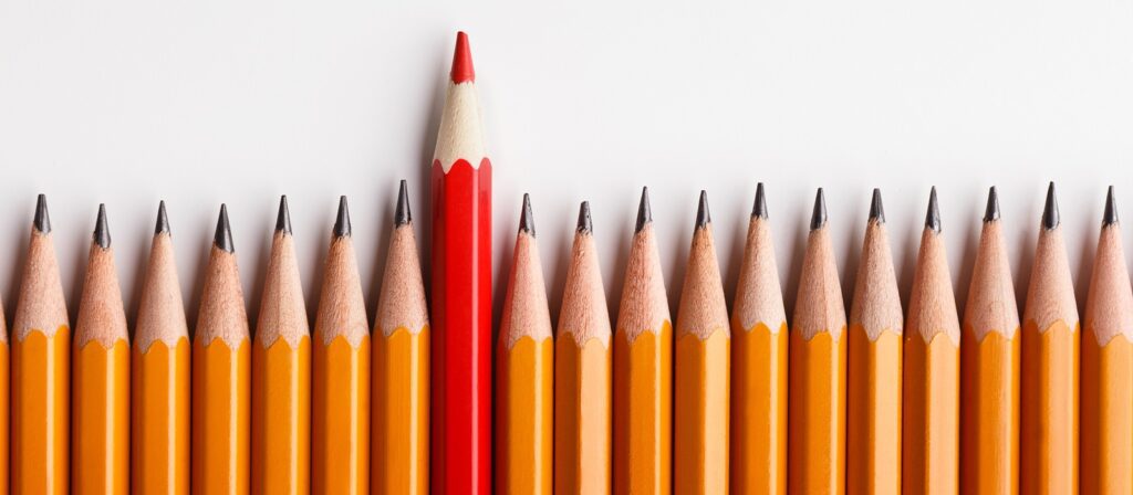 A row of yellow pencils lined up side by side, with one red pencil standing upright among them, contrasting against the white background.