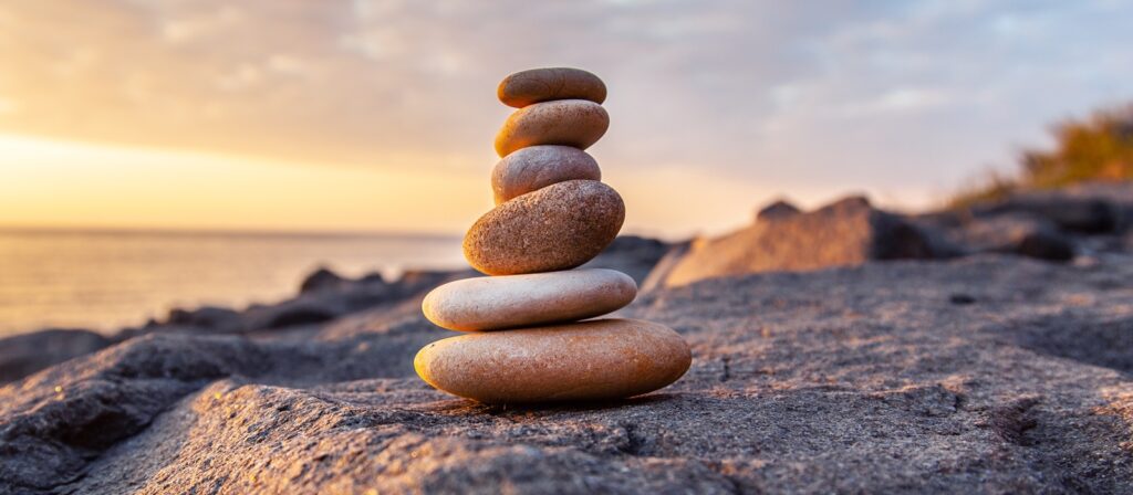 A stack of smooth, flat stones balanced on a rocky surface near a body of water at sunset, with soft golden light and a blurred background.