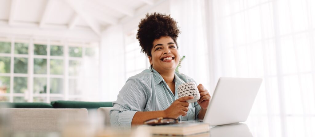 A smiling woman sits at a table with a laptop and holds a patterned mug. She appears relaxed and happy in a bright room with large windows and soft natural light.