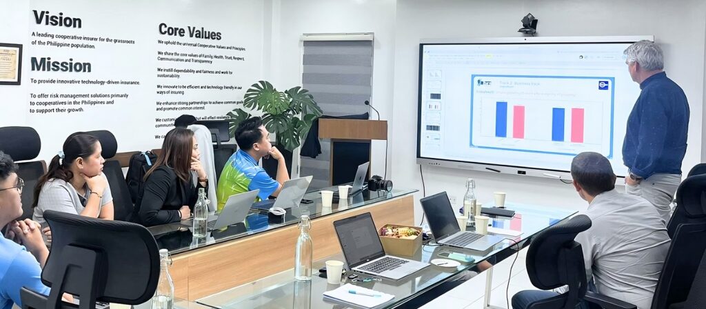 A group of people in a meeting room watch a man present bar graphs on a large screen. Laptops, drinks, and snacks are on the table. Vision, Mission, and Core Values are displayed on the wall.