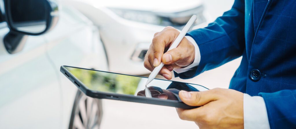 A person in a blue suit uses a stylus to write on a tablet, with blurred cars visible in the background.