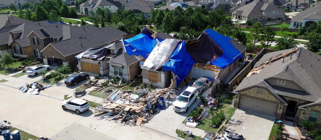 A neighborhood with several homes; one house is heavily damaged, covered in blue tarps, with debris and construction materials scattered in front, and cars parked along the street.