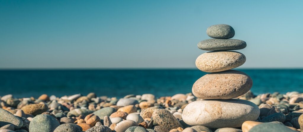 A stack of five smooth stones balanced on a pebble-covered beach, with a calm blue sea and clear sky in the background.