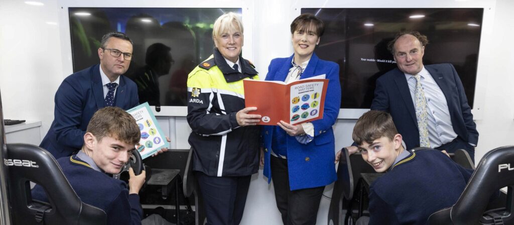 A group of adults and teenagers pose indoors; two adults hold books and wear uniforms, while three teens sit at gaming simulators, all smiling at the camera. Monitors and equipment appear in the background.