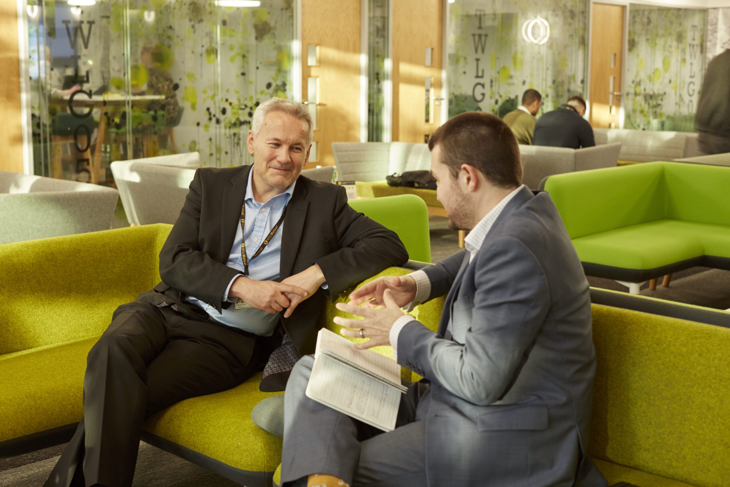Two men in business attire sit and talk on green sofas in a modern office lounge. One man listens while the other gestures with his hands over an open notebook. Others work in the background.