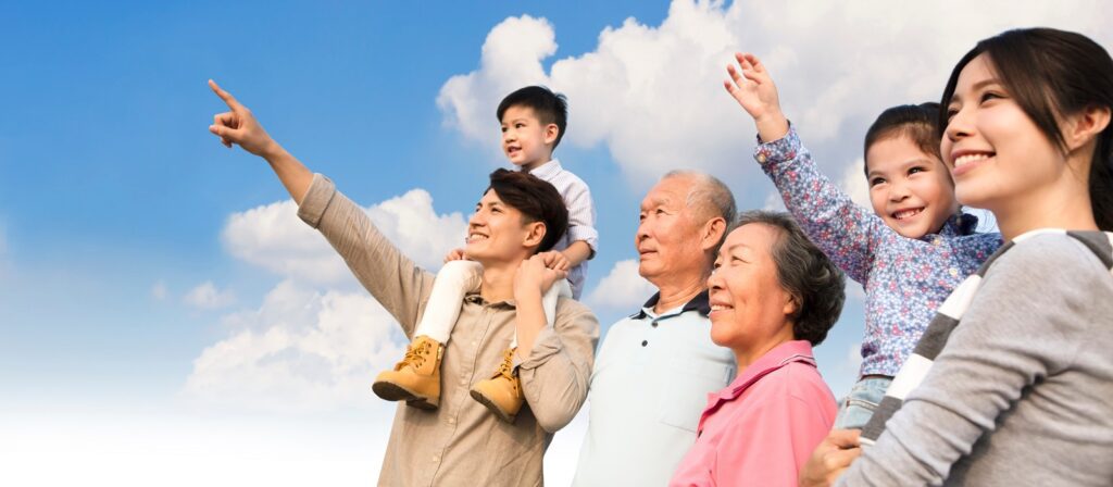 A multigenerational Asian family stands outdoors under a blue sky with clouds. A man points ahead while carrying a child on his shoulders. The family smiles and looks in the same direction, appearing happy and united.