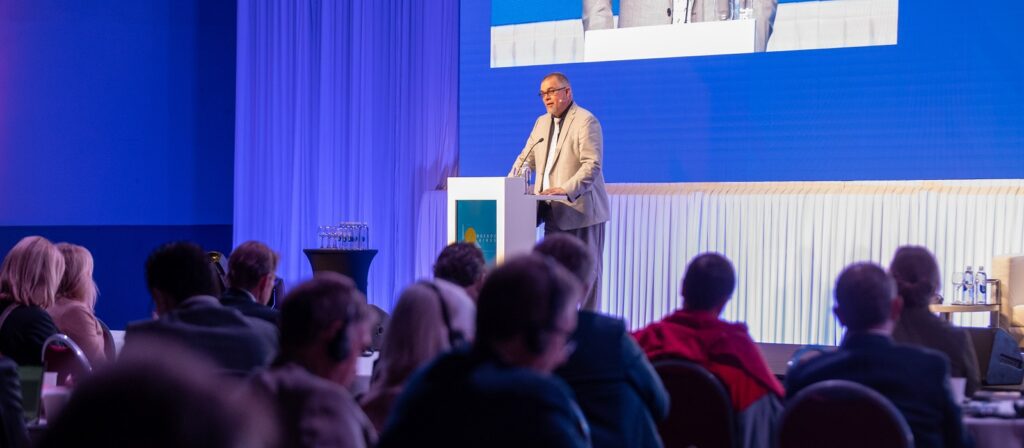 A man in a light-colored suit speaks at a podium on a stage in front of an audience. Behind him is a large blue screen displaying his image. The audience is seated at round tables.