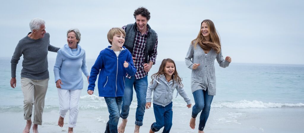 Three adults, two children, and an elderly couple laugh and walk barefoot along the beach, dressed in casual, warm clothing on a cloudy day by the sea.