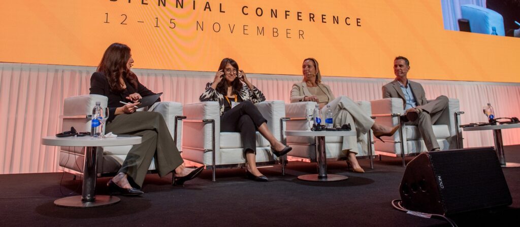 Four people sit on stage in white chairs during a panel discussion at a conference. Behind them, an orange screen displays event details. Tables with water bottles are in front of the panelists.