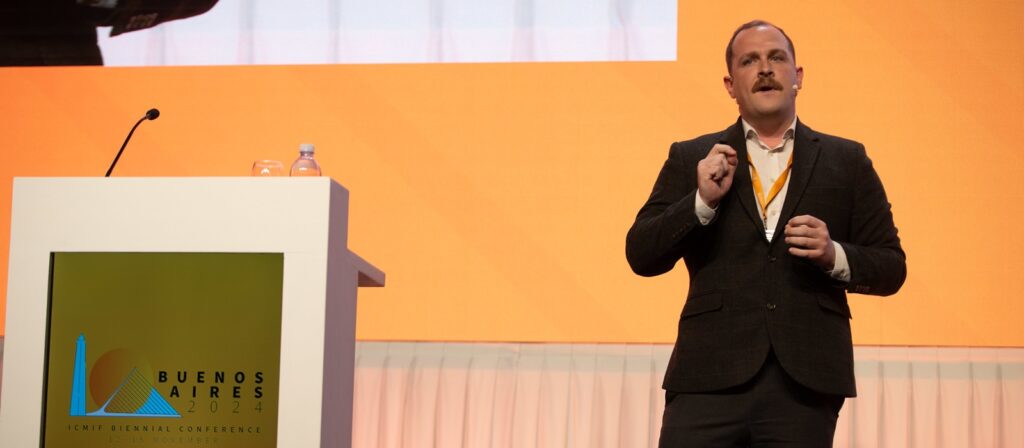 A man in a suit speaks on stage next to a podium with the “Buenos Aires 2024 ICMF Biennial Conference” logo, against an orange background. He wears a microphone headset and gestures with his hand.