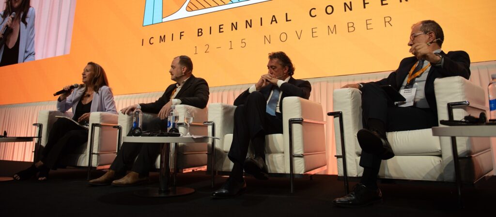 Four people sit on a stage in white chairs at the ICMIF Biennial Conference, with microphones and water bottles on a table. An orange screen behind them displays the conference name and dates: 12-15 November.