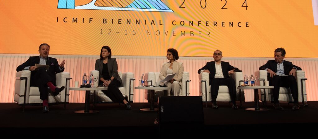 Five people sit onstage in armchairs during a panel discussion at the ICMIF Biennial Conference 2024. The backdrop is orange with conference details and dates, and each person has water bottles and microphones nearby.