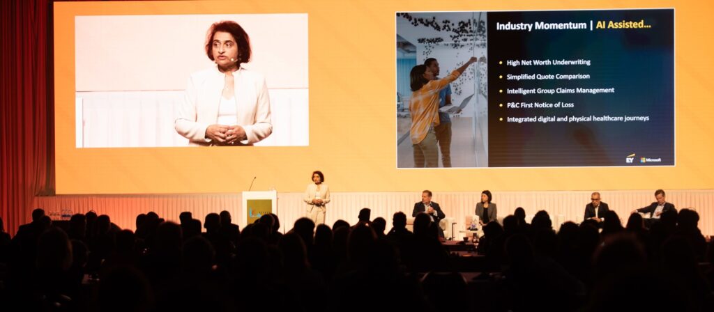 A woman stands on stage presenting with a large screen behind her showing Industry Momentum | AI Assisted... and a list of topics, while four panelists sit in the background and audience members watch.
