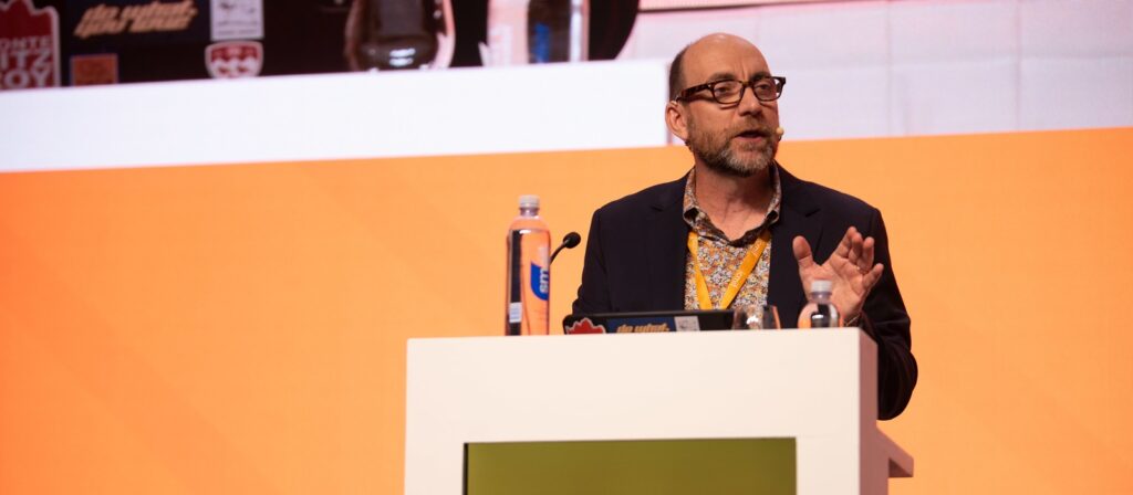 A man with glasses and a beard is speaking on stage at a podium with a microphone, water bottles, and a laptop, against an orange and white background.