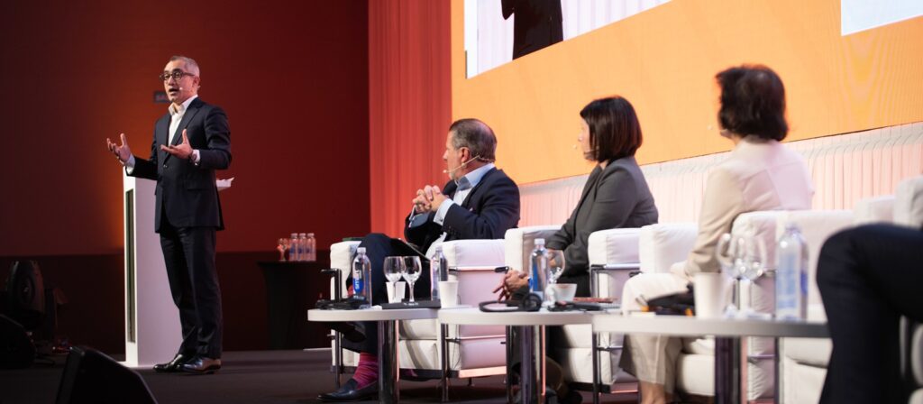 A man in a suit speaks at a podium on stage, while three people sit on a panel with microphones, listening. Bottled water and glasses are on a table in front of them; an orange and white screen is in the background.