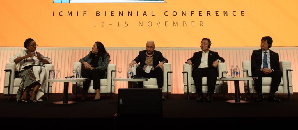 Five people sit on stage in armchairs during a panel discussion at the ICMIF Biennial Conference, with microphones and water bottles on tables in front of them and a large orange backdrop displaying the event details.