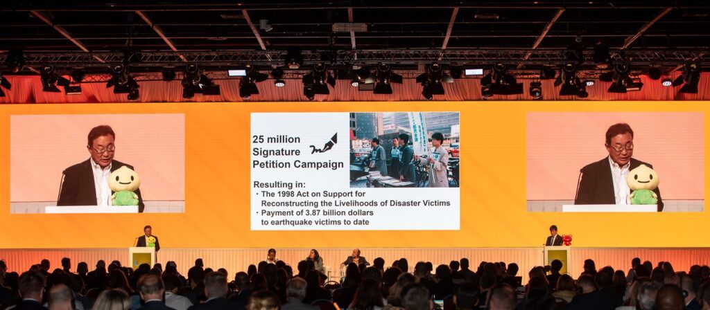 A speaker stands at a podium addressing an audience. Behind them, a large screen displays information on a signature petition campaign for disaster victims, including images, text, and a plush toy on the podium.