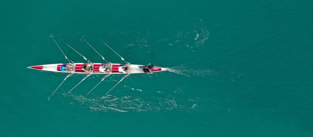 Aerial view of four people rowing in a narrow boat on clear turquoise water, leaving ripples behind as they move forward in unison.