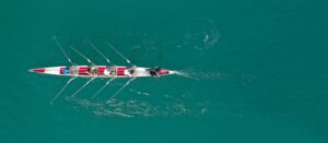 Aerial view of four people rowing in a narrow boat on clear turquoise water, leaving ripples behind as they move forward in unison.