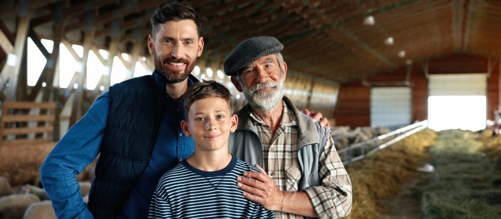 Three generations of men—an elderly man, a middle-aged man, and a young boy—stand close together inside a barn, smiling at the camera with sheep and hay visible in the background.