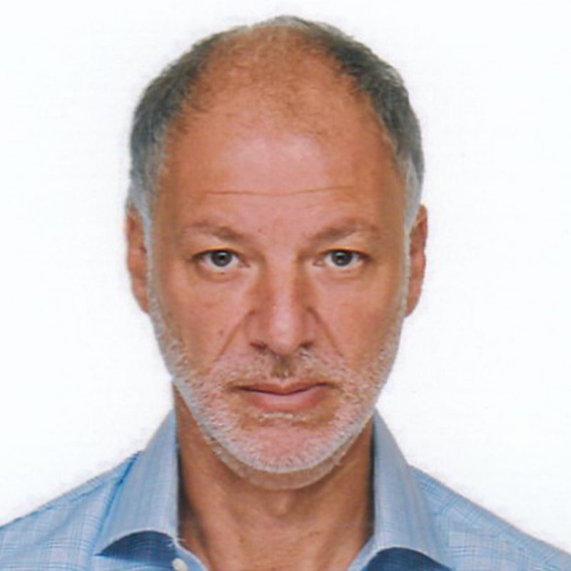 Portrait of a middle-aged man with short gray hair, light skin, and a trimmed gray beard, wearing a light blue collared shirt, looking directly at the camera against a plain white background.