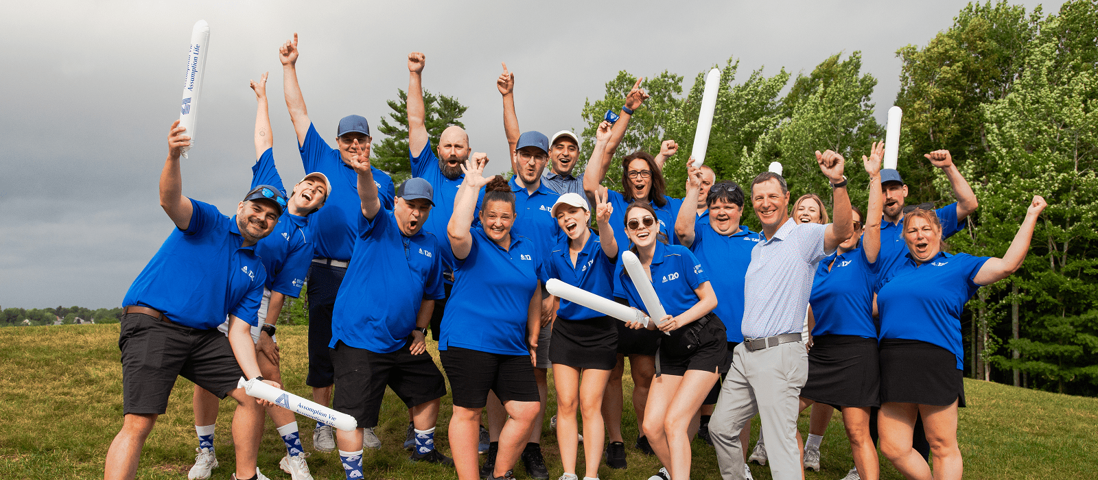 A group of people in matching blue shirts and caps cheer enthusiastically outdoors, holding inflatable noise sticks, with trees and a cloudy sky in the background.