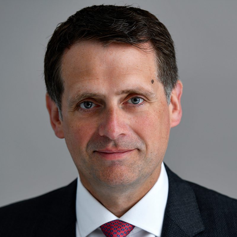A man with short dark hair wearing a dark suit, white shirt, and red patterned tie, looking directly at the camera with a neutral expression against a plain gray background.