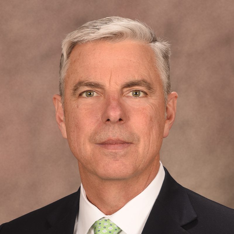 A middle-aged man with short gray hair wearing a dark suit, white shirt, and light green patterned tie, posing in front of a plain brown background.