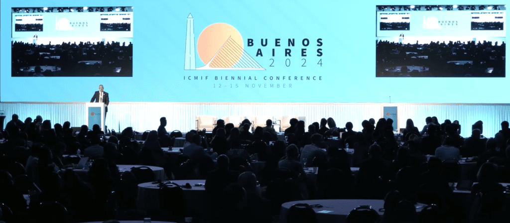 A speaker stands on stage addressing an audience at the ICMIF Biennial Conference 2024 in Buenos Aires, with a large blue backdrop displaying the event details and logo.