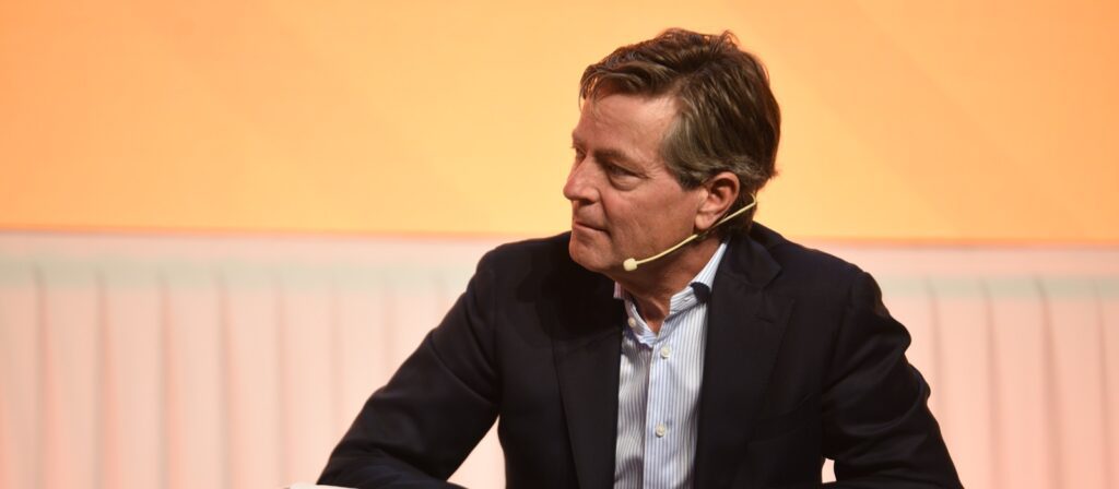 A man in a dark suit and white shirt sits, looking to the side, wearing a headset microphone, with a softly lit orange and white background behind him.