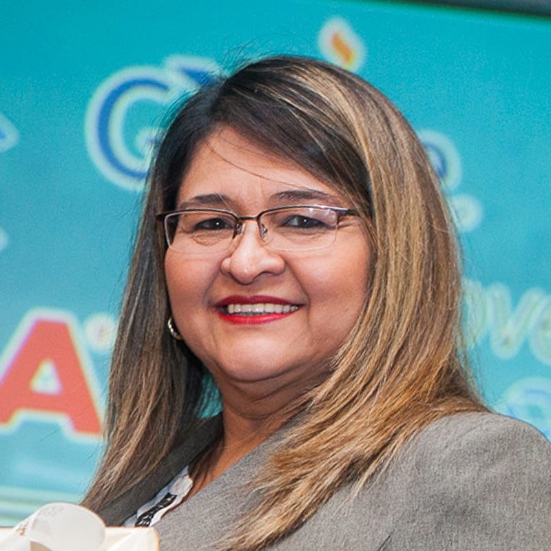 A woman with straight, shoulder-length brown hair and glasses smiles at the camera. She is wearing a gray blazer and standing in front of a blue background with colorful text and shapes.