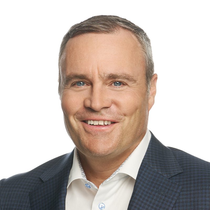 A smiling man with short gray hair wearing a dark checked suit jacket and a white collared shirt, posed against a plain white background.