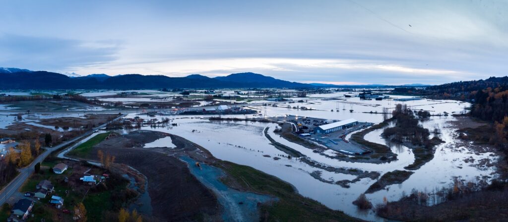 Aerial view of a flooded rural landscape with submerged fields, roads, and buildings; water covers much of the area, surrounded by trees and distant mountains under a cloudy sky.