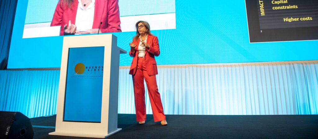 A woman in a red suit speaks on stage at a conference in Buenos Aires, standing behind a podium, with a large blue screen and presentation slides displayed in the background.