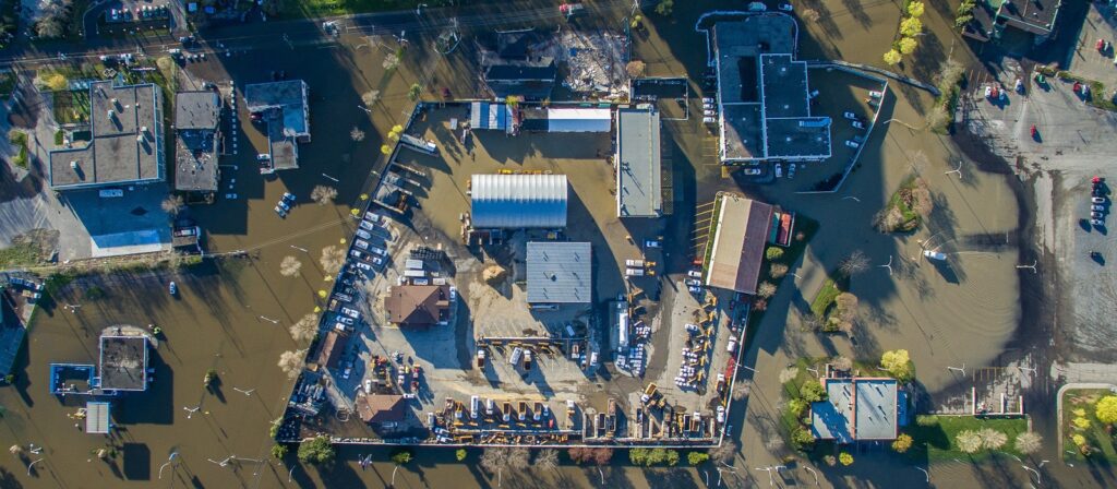 Aerial view of a flooded industrial area surrounded by muddy water, with partially submerged buildings, vehicles, and equipment visible inside and around the fenced perimeter. Trees and rooftops protrude from the floodwater.