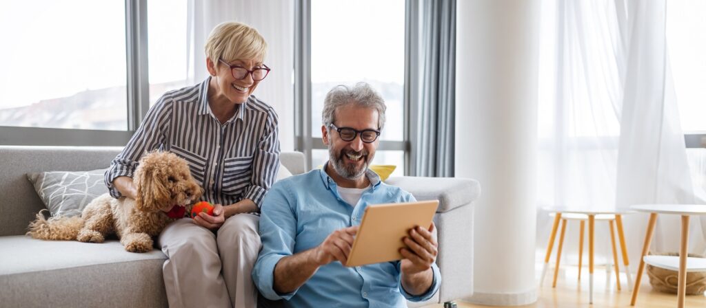 An older couple sits in a bright living room; the woman pets a small brown dog on the couch while the man, sitting on the floor, smiles at a tablet. Sunlight streams through large windows behind them.