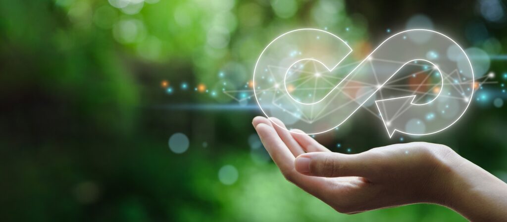 A hand holds a glowing, transparent infinity loop with arrows, symbolizing sustainability or circular economy, against a blurred green natural background.