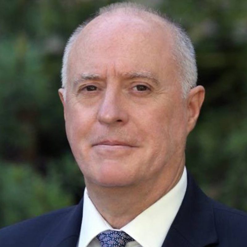 A middle-aged man with short gray hair is wearing a dark suit, white shirt, and patterned tie. He is outdoors, standing in front of greenery, and looking directly at the camera with a neutral expression.