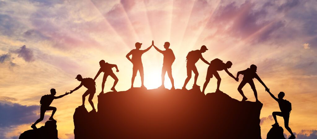Silhouettes of people helping each other climb a mountain at sunset, with sun rays shining in the background, symbolizing teamwork, support, and achievement.
