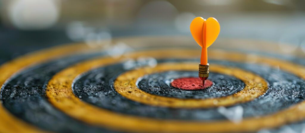 A yellow dart with a heart-shaped tail hits the red bullseye at the center of a black and yellow dartboard.