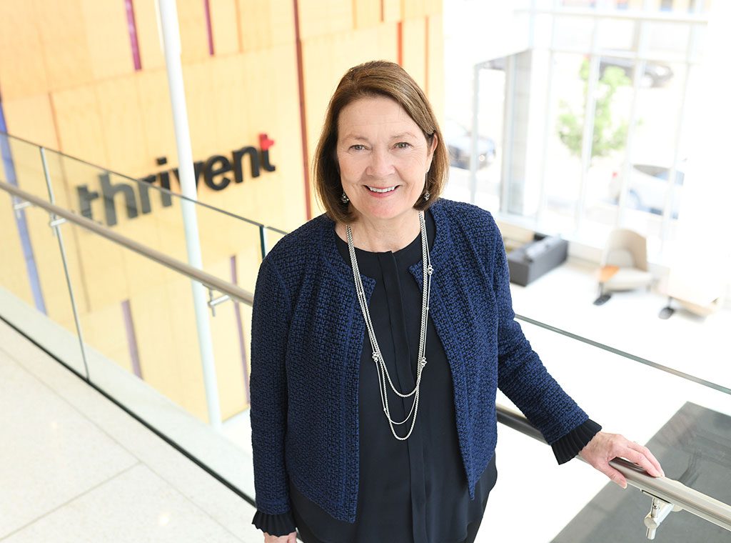 A woman with shoulder-length brown hair, wearing a navy blazer and layered necklace, smiles while standing indoors near a glass railing. The word thrivent is displayed on a wall in the background.